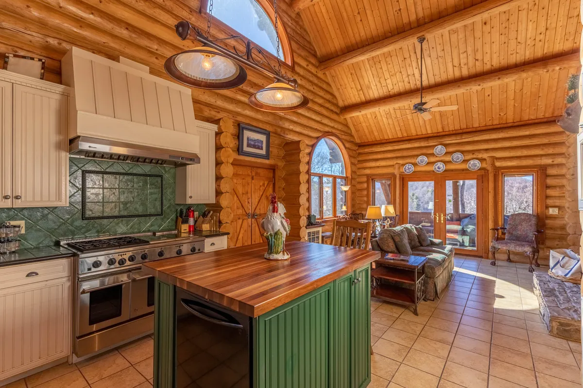 A wider kitchen/dining area view that captures the kitchen island, log walls, large windows, and an adjacent dining space. The natural light and decorative touches create a lively atmosphere, emphasizing the transition between kitchen activities and shared meals.