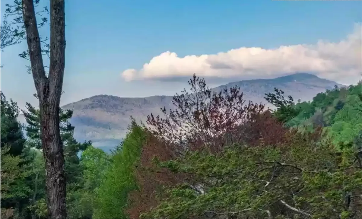 A panoramic view of the surrounding mountain range with clouds drifting above and trees in the foreground. The tranquil landscape sets the scene for hiking, nature walks, and photography, all core to the Blowing Rock adventure theme.