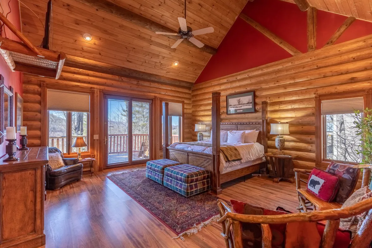 A warmly decorated bedroom featuring a four-poster bed, large windows that allow natural light to stream in, a cozy seating area, and a wooden dresser. The space feels inviting and provides both comfort and privacy, with scenic views to enjoy from bed or the reading nook.