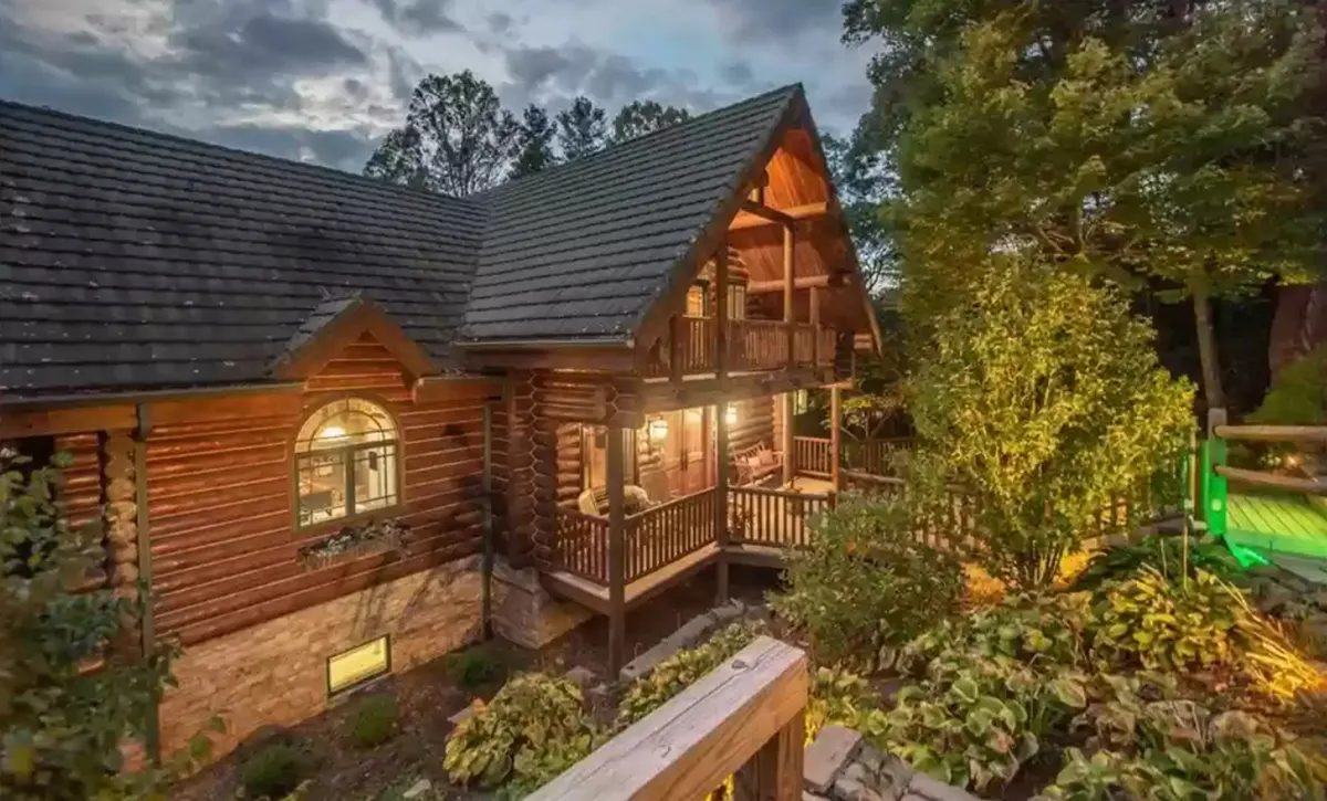 This exterior image shows the log cabin structure nestled among lush greenery, with a balcony featuring inviting seating and warm outdoor lighting. The scene radiates a cozy, welcoming atmosphere and hints at the adventure-ready setting, with outdoor spaces ideal for relaxing after a day of exploring.