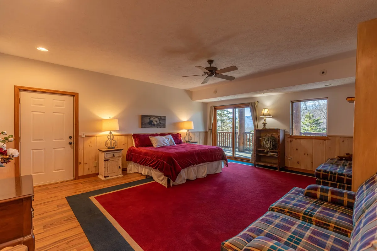 A bedroom scene featuring a large bed with red bedding and patio doors leading to a balcony, suggesting an easy transition from indoor relaxation to outdoor exploration. The doors open up to the fresh mountain air and potential scenic views, further tying the interior to the broader natural context.