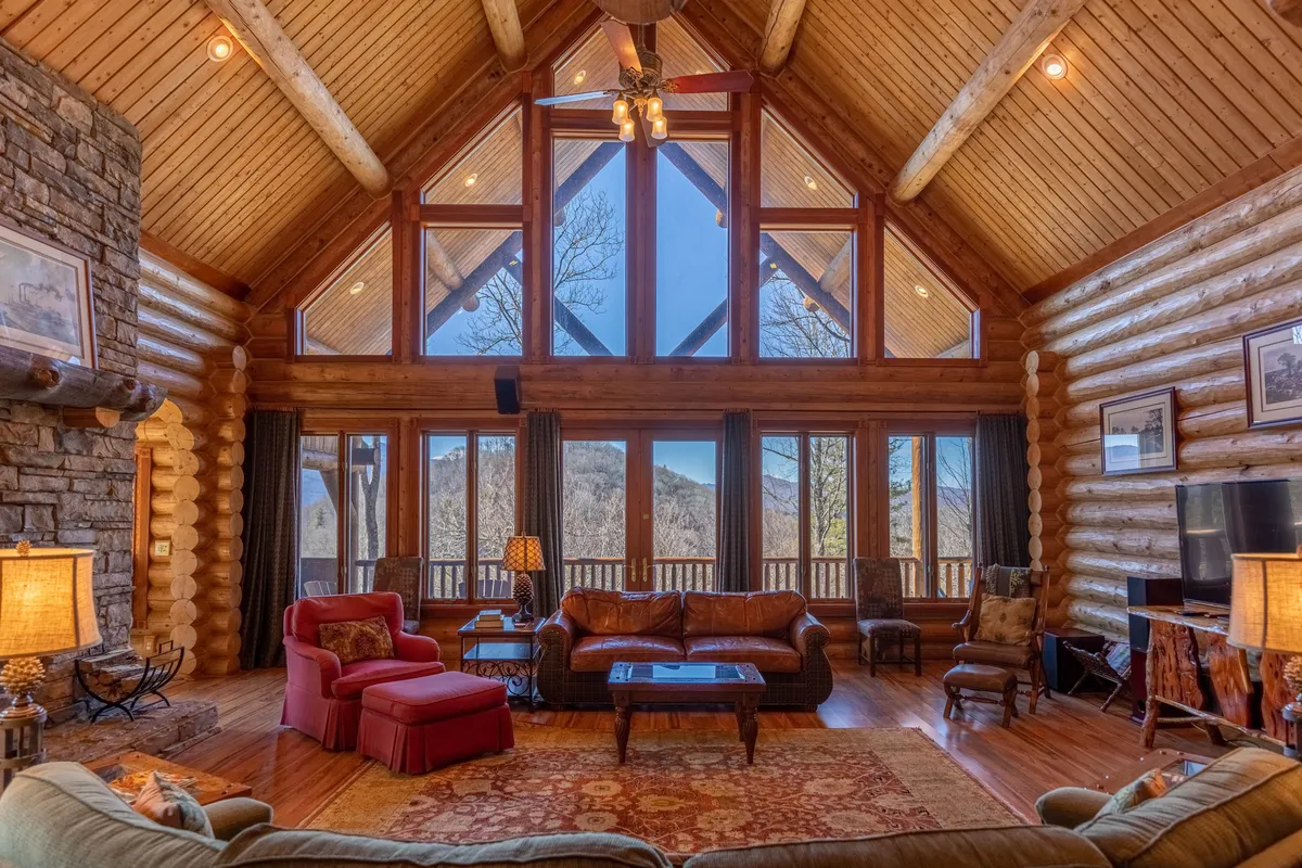 This image captures a spacious interior living area featuring large windows with panoramic mountain views, a statement stone fireplace, exposed wooden beams, log accents, and plush, comfortable seating arranged for conversation and relaxation. Natural light pours in, creating an inviting setting that highlights the cabin's luxury and rustic charm.