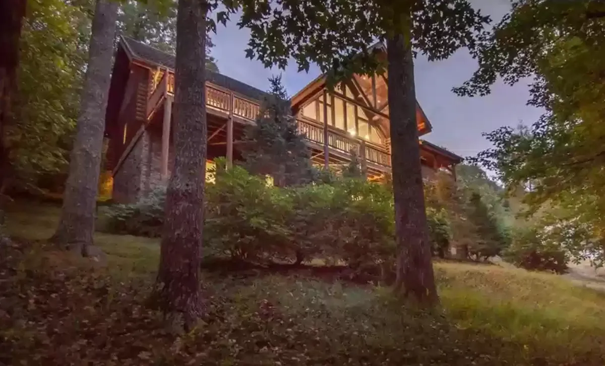 The image highlights the striking wooden log cabin set among lush trees, with a spacious balcony bordered by rustic railings. The cabin's elevated position offers hints of sweeping mountain views, and the inviting balcony suggests space for family gatherings or tranquil evenings. There's a welcoming, peaceful atmosphere that showcases the property's blend of luxury and nature.