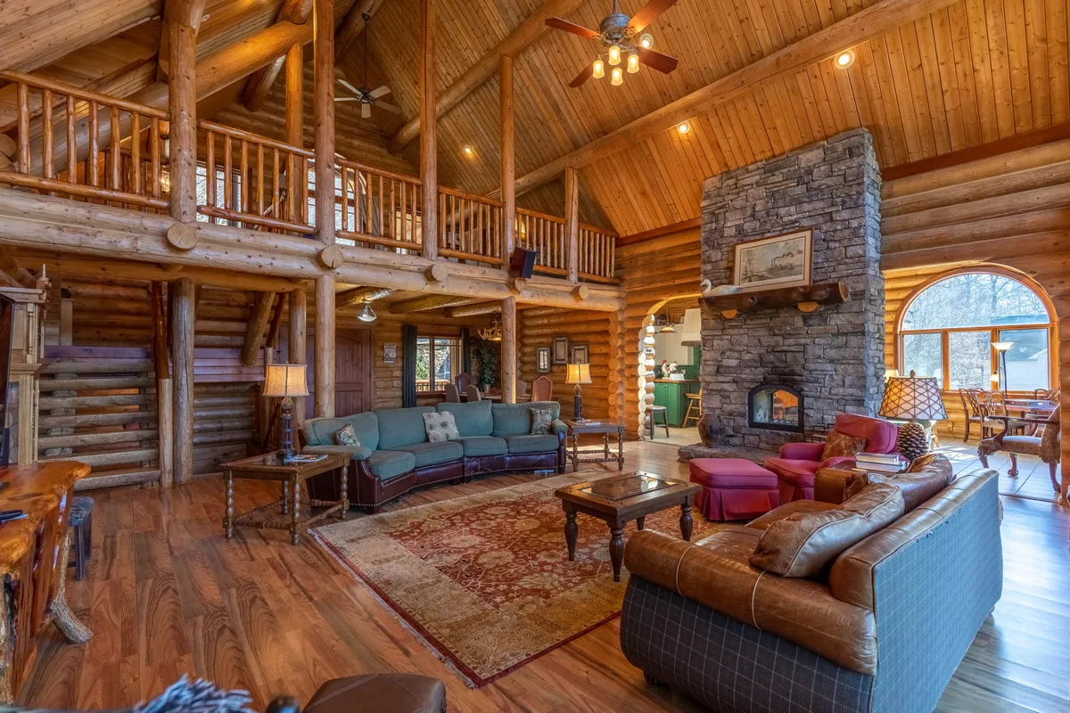 A large, open interior living space highlighted by a stunning stone fireplace, expansive windows letting in natural light, exposed wooden beams, and comfortable seating. The room is both cozy and spacious, ideal for family gatherings or relaxing after a mountain adventure.
