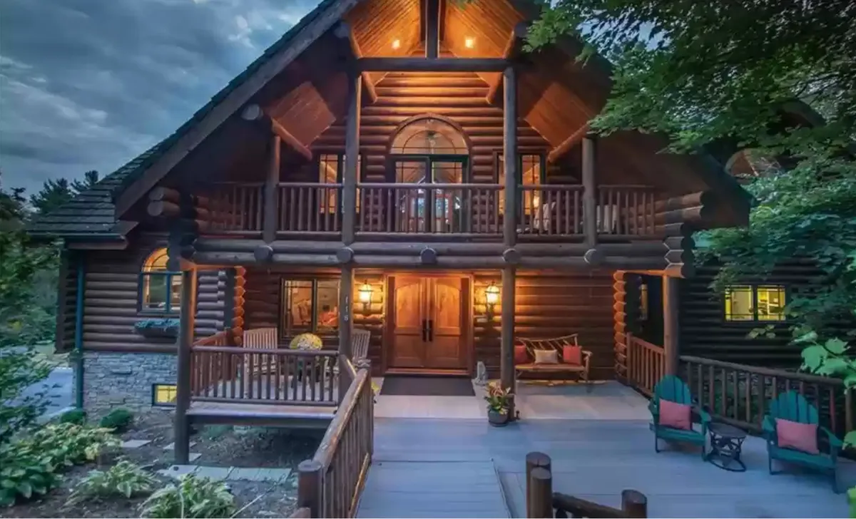This image showcases the exterior of Bear Lodge, featuring its classic log cabin architecture, a welcoming front porch illuminated in the evening, a balcony above, and a landscaped garden. The scene is set for gatherings, with inviting spaces perfect for family moments or relaxing after a day of adventure.