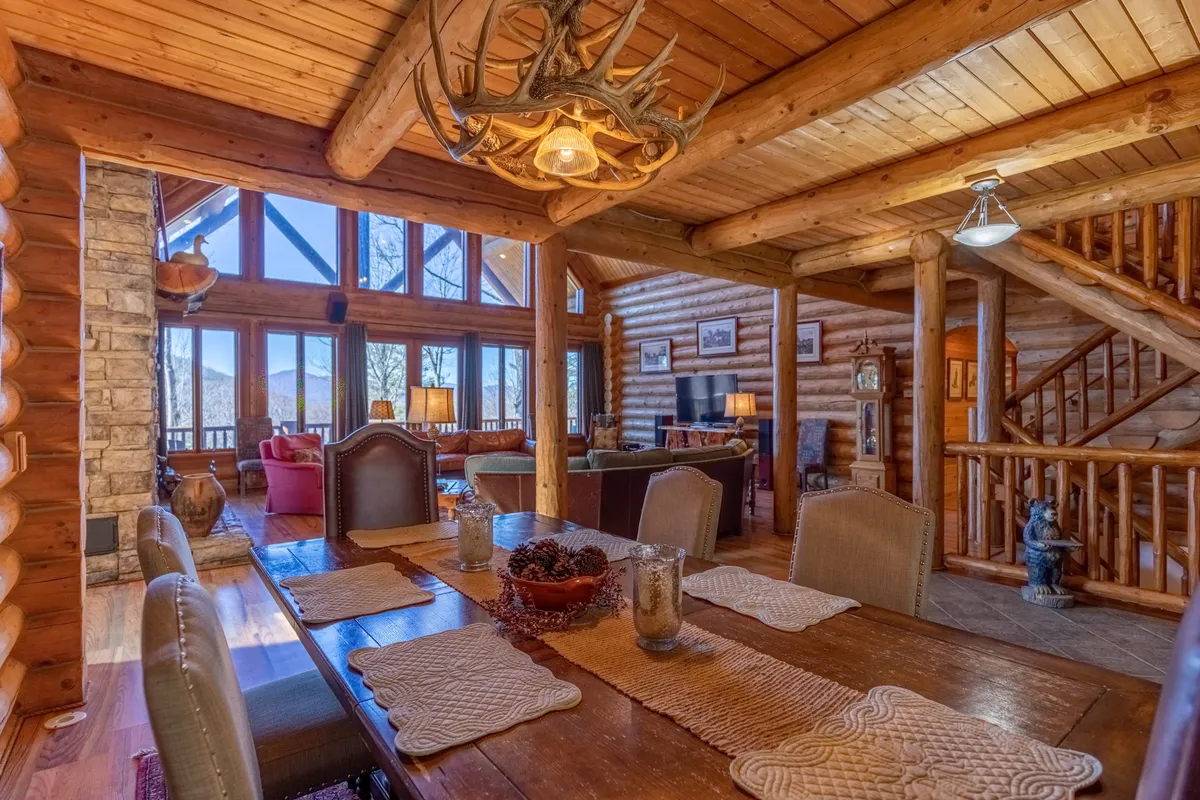 A rustic dining room scene with a large wooden table, antler chandelier, stone fireplace, and expansive windows revealing breathtaking mountain views. The arrangement invites group dining and holiday celebrations in an atmosphere full of warmth and natural beauty.