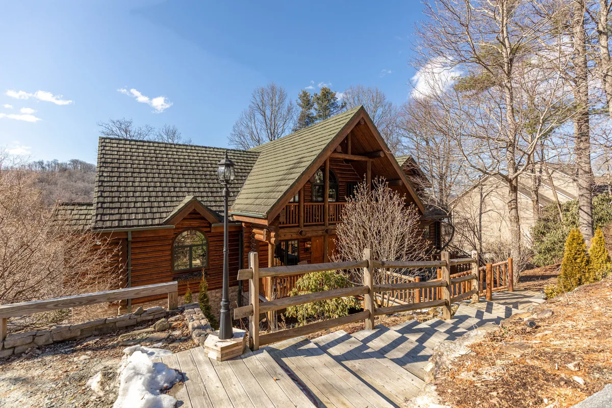 A cozy log cabin with classic architecture sits nestled in nature, featuring a wooden walkway and spacious porch. The surrounding greenery and inviting porch evoke a sense of rustic charm and connection to the outdoors, suggesting opportunities for nature walks and relaxing moments outside.