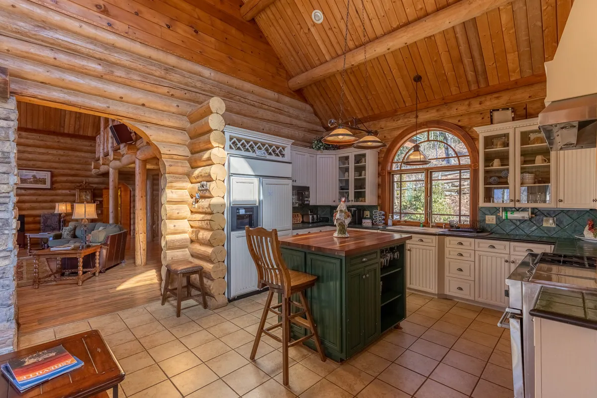 A warm, inviting kitchen space featuring log walls, exposed wooden beams, a large window drawing in natural light, and a spacious kitchen island at the center. The view includes ample counter space and the island, ideal for group meal prep or casual gatherings. The open layout hints at easy flow into adjacent living and dining areas.