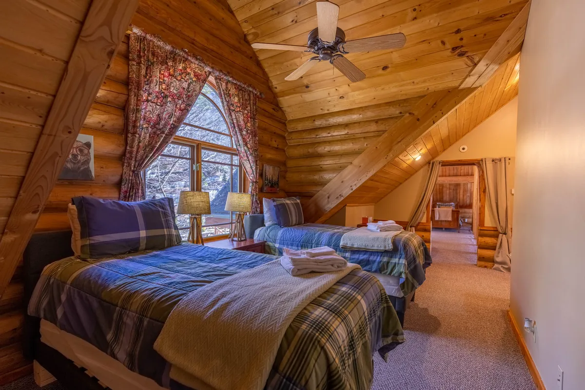 A bedroom designed for flexibility and comfort, featuring two twin beds with plaid bedding, a large window offering views of the outdoors, wooden beams, and colorful curtains. The room is bright and airy, ideal for children or extra guests as a flexible sleeping area.