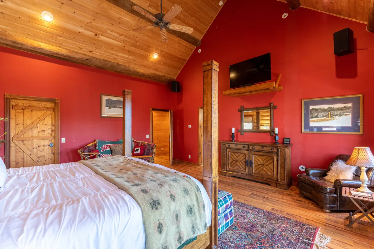 A spacious bedroom with a large bed dressed in a decorative blanket, wooden furniture, a wall-mounted TV, vibrant red walls, and a cozy seating area by the window. The atmosphere is warm and inviting, perfect for unwinding after a busy day.